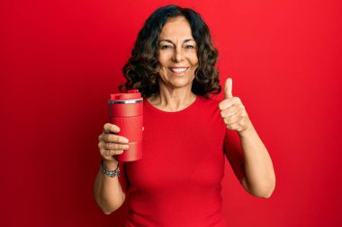 Orta yaşlı İspanyol kadın bir fincan kahve içiyor mutlu ve pozitif gülümsüyor, başparmağıyla mükemmel ve onay işareti yapıyor. 