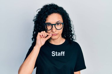 Kıvırcık saçlı, personel tişörtü giyen ağzı ve dudakları parmaklarıyla kapanan İspanyol bir kadın. gizli ve sessiz, tabu konuşması 