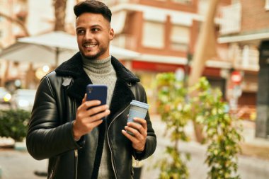 Genç Arap adam akıllı telefon kullanıyor ve şehirde kahve içiyor..