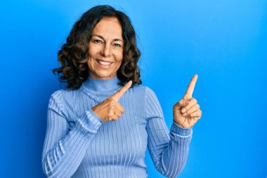 Orta yaşlı İspanyol kadın günlük kıyafetler giyiyor gülümsüyor ve kameraya bakıyor iki eliyle ve parmaklarıyla kenarı işaret ediyor.. 