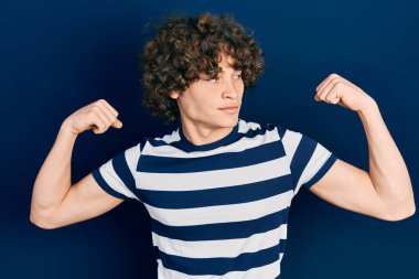 Sıradan çizgili tişört giyen yakışıklı genç adam gururla gülümseyen kol kaslarını gösteriyor. fitness konsepti. 