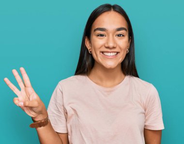 Genç Asyalı kadın rahat kıyafetler giyip üç numaralı parmaklarıyla işaret ederken kendinden emin ve mutlu bir şekilde gülümsüyor.. 