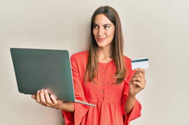 Genç ve güzel bir kadın dizüstü bilgisayar ve kredi kartı tutuyor gülümsüyor ve yan bakıp düşünüyor.. 