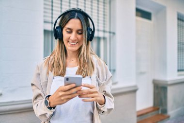 Beyaz kadın şehirde akıllı telefon ve kulaklık kullanarak mutlu bir şekilde gülümsüyor..
