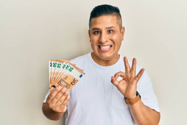Elinde 50 avroluk banknotlar tutan genç Latin adam parmaklarıyla imza atıyor, gülümseyerek mükemmel bir sembol oluşturuyor. 