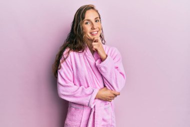 Bornoz giyen genç sarışın kadın kameraya kendinden emin bakıyor kollarını kavuşturup gülümseyerek ve çenesini kaldırarak. Olumlu düşünmek. 