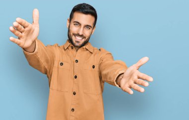 Gündelik giysiler giymiş genç İspanyol adam kameraya bakıp sarılmak için kollarını açmış gülümsüyor. Mutluluğu kucaklayan neşeli bir ifade.. 