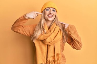 Genç beyaz kadın yün kazak ve kep giyip neşeli gülüyor ve parmakları ve ağzı ile işaret ediyor. diş sağlığı kavramı. 