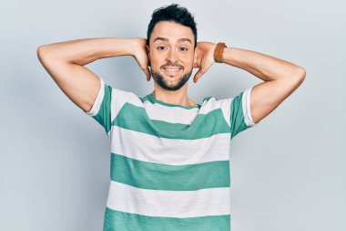 Genç İspanyol bir adam günlük kıyafetler giyiyor rahatlıyor ve esniyor, kolları ve elleri başının arkasında mutlu bir şekilde gülümsüyor. 