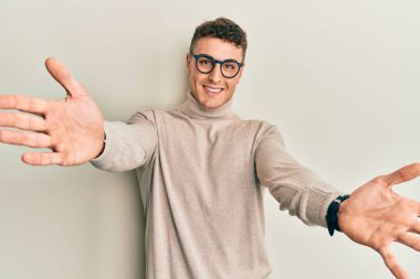 Balıkçı yaka kazak giyen İspanyol genç adam sarılmak için kollarını açmış gülümseyen kameraya bakıyor. Mutluluğu kucaklayan neşeli bir ifade.. 