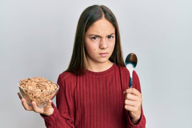 Güzel esmer küçük kız sağlıklı tahıl gevrekleri yiyor kaşıkta şüpheci ve gergin tutuyorlar sorun yüzünden surat asıyorlar. negatif kişi. 