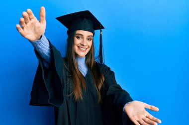 Mezuniyet şapkası ve tören cübbesi giymiş güzel esmer kadın kucak açmak için kollarını açıp kameraya bakıyor. Mutluluğu kucaklayan neşeli bir ifade.. 