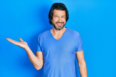 Orta yaşlı, günlük kıyafetler giyen, neşeli gülümseyen, elinde avucuyla kameraya bakan beyaz bir adam.. 