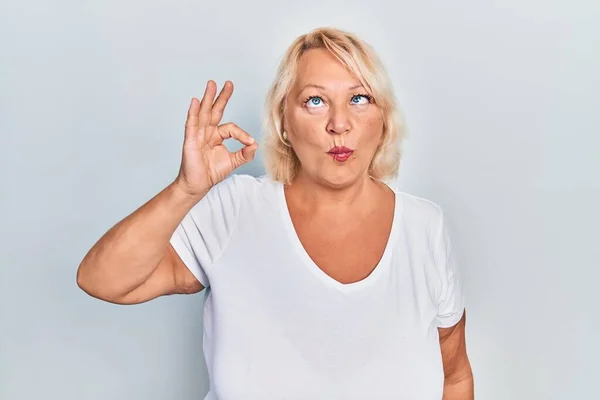 Middle age blonde woman doing ok positive gesture making fish face with mouth and squinting eyes, crazy and comical.