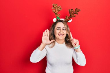 Orta yaşlı beyaz kadın şirin Noel geyiği boynuzları takıyor. Altıncı parmaklarını gösterip gösterirken kendinden emin ve mutlu bir şekilde gülümsüyor.. 