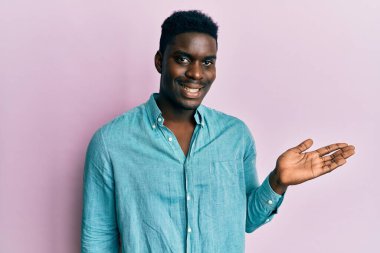 Yakışıklı, sıradan kıyafetler giyen, neşeli bir şekilde gülümseyen, avuç içiyle kameraya bakan siyah bir adam.. 