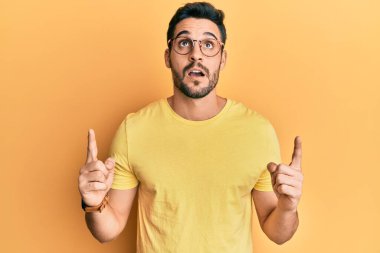 Gündelik giysiler ve gözlük takan genç İspanyol adam şaşırmış ve şaşırmış bir şekilde yukarı baktı ve parmaklarıyla işaret etti ve kollarını kaldırdı.. 