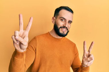 Sakallı, sıradan bir kazak giyen genç adam kameraya bakıp zafer işareti yapıyor. İki numara.. 