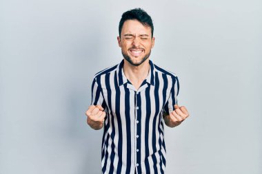 Gündelik kıyafetler giyen genç İspanyol adam çok mutlu ve heyecanlı. Kazanan jestler yapıyor. Kolları havada, gülümsüyor ve başarı için bağırıyor. kutlama konsepti. 