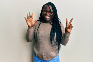 Saçları örgülü, günlük kıyafetler giyen ve gözlük takan genç siyahi bir kadın yedi numaralı parmaklarıyla gülümseyerek mutlu ve kendinden emin bir şekilde onu işaret ediyor.. 