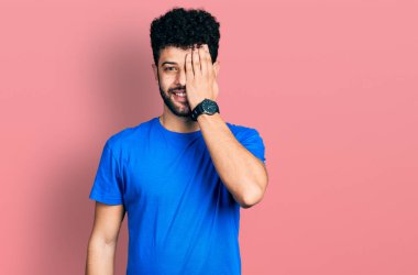 Sakallı genç, mavi tişörtlü bir Arap bir gözünü eliyle kapatıyor, yüzünde kendine güvenen bir gülümseme ve sürpriz bir duygu var.. 