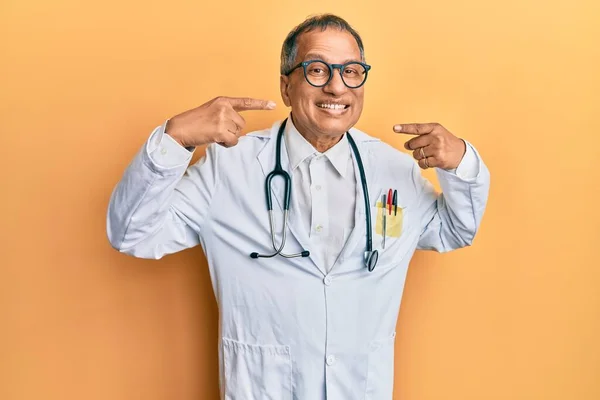 医者のコートと聴診器を身に着けている中年のインド人男性は 明るい表情と指の歯と口で指して笑っています 歯の健康の概念 — ストック写真