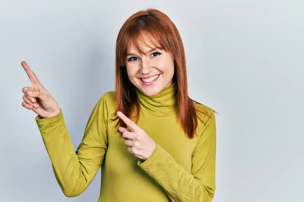红头发的年轻女人穿着高领毛衣 微笑着看着摄像机 用双手和手指指向旁边 — 图库照片