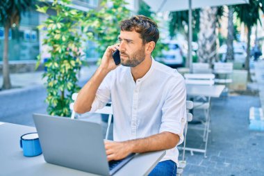 Güneşli bir günde günlük beyaz gömlek giyen sakallı yakışıklı adam laptopla çalışıyor ve kafeteryada telefonla konuşuyor.