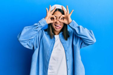 Tatlı yün şapkalı genç Latin kadın dürbün gibi jest yapıyor, gözleri parmaklarından bakıyor. çılgın ifade. 