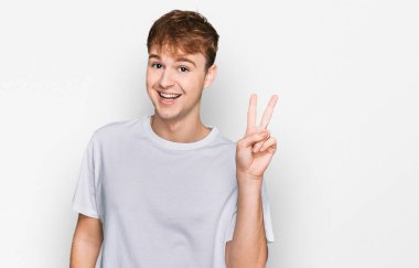 Beyaz günlük tişört giyen genç beyaz bir adam iki numarayı gösterip işaret ederken kendinden emin ve mutlu gülümsüyor.. 