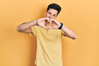 Gündelik sarı tişört giyen, aşık gülümseyen, elleriyle kalp sembolü yapan genç İspanyol adam. romantik kavram. 
