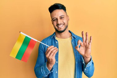 Elinde Litvanya bayrağı tutan genç Arap adam parmaklarıyla işaret veriyor, gülümseyerek mükemmel bir sembol oluşturuyor. 