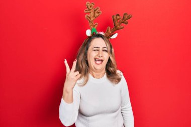 Orta yaşlı, beyaz bir kadın. Geyik boynuzları takıyor. Kameraya gülümsüyor ve zafer işareti yapıyor. İki numara.. 