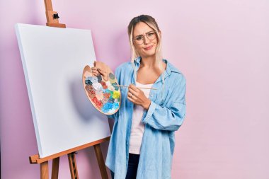 Güzel, genç, sarışın bir kadın boya sehpası ile resim çiziyor şüpheci ve gergin duruyor, sorun yüzünden surat asıyor. negatif kişi. 