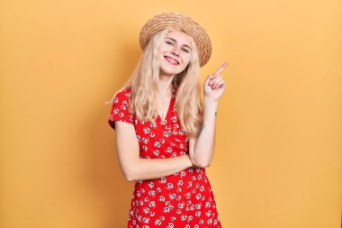 Sarışın, yaz şapkası takan, yüzünde kocaman bir gülümseme olan, eli ve parmağı kameraya bakan güzel beyaz bir kadın.. 