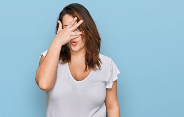 Genç ve cüsseli bir kadın günlük beyaz tişört giyiyor. Şok olmuş bir şekilde yüzünü ve gözlerini kapatıyor. Utanmış bir ifadeyle parmaklarının arasından bakıyor.. 