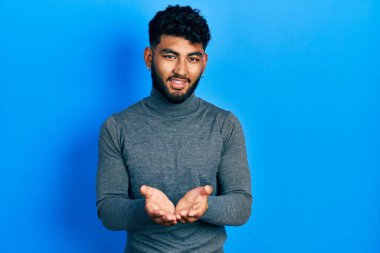 Balıkçı yaka kazak giyen ve elleri avuçlarında gülümseyen sakallı bir Arap, jest yapıyor. Bekleme ve koruma 