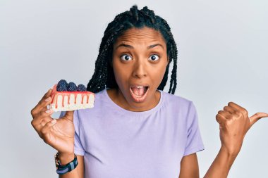 Saçları örgülü Afrikalı Amerikalı bir kadın bir dilim cheesecake tutuyor başparmağını yana uzatıyor ve ağzı açık gülümsüyor. 