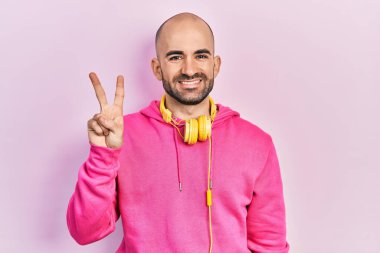 Spor kıyafetleri giyen, kulaklık takan genç kel bir adam iki numarayı gösterip işaret ederken kendinden emin ve mutlu bir şekilde gülümsüyor.. 