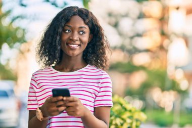 Genç Afrikalı Amerikalı kız şehirde akıllı telefon kullanarak mutlu bir şekilde gülümsüyor..