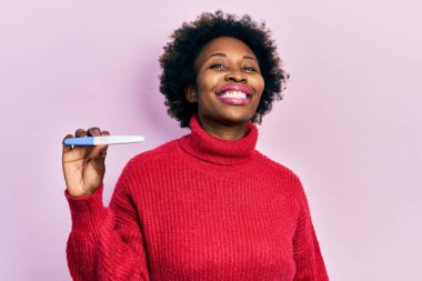 Termometreyi tutan Afro-Amerikalı genç bir kadın pozitif ve mutlu görünüyor ve kendine güvenen bir gülümsemeyle dişlerini gösteriyor. 