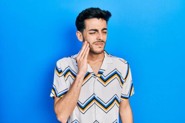 Günlük kıyafetler giyen genç İspanyol adam diş ağrısı ya da diş hastalığından dolayı acı dolu bir ifadeyle ağıza dokunuyor. dişçi 