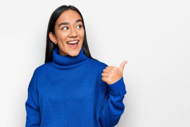 Genç Asyalı kadın, günlük kışlık süveter giyiyor, gülümsüyor, gülümsüyor ve başparmağıyla yan tarafa bakıyor.. 