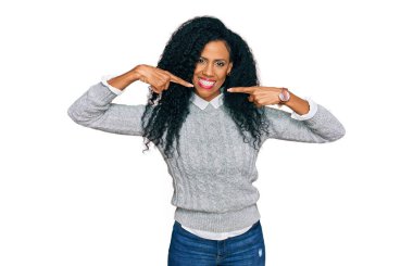 Orta yaşlı Afro-Amerikalı kadın gündelik kıyafetler giyip gülümsüyor ve parmakları, dişleri ve ağzı ile işaret ediyor. diş sağlığı kavramı. 