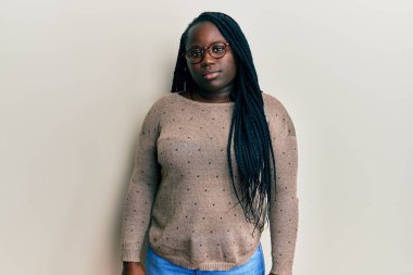 Saçları örgülü, günlük kıyafetler giyen ve gözlüklü genç siyahi bir kadın. Yüzünde ciddi bir ifadeyle rahatlamış. Basit ve doğal bir şekilde kameraya bakıyorsun.. 