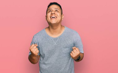 Gündelik kıyafetler giyen genç latin adam çok mutlu ve heyecanlı. Kolları havada, gülümseyen ve başarı için çığlık atan kazanan bir jest yapıyor. kutlama konsepti. 