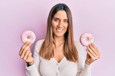 Güzel pembe çörekler tutan esmer bir kadın yüzünde mutlu ve serin bir gülümsemeyle gülümsüyor. diş gösterme. 