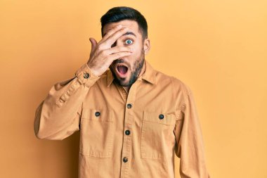 Gündelik kıyafetler giyen genç İspanyol adam şok içinde yüzünü ve gözlerini eliyle kapatıyor, parmaklarından korkuyla bakıyor. 