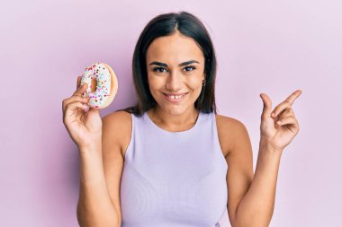 Genç esmer kadın elinde lezzetli renkli donut tutarken gülümsüyor mutlu bir şekilde elini ve parmağını yana uzatıyordu. 