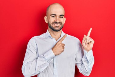 Zarif kıyafetler giymiş genç kel bir adam gülümseyip kameraya bakıyor ve iki eliyle ve parmaklarıyla kenarı işaret ediyor.. 